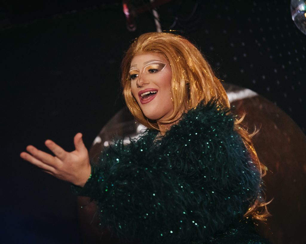Promotional image of a stage performer wearing a glittering dark green feathered costume and a long blonde wig, illuminated by warm stage lighting against a dark backdrop.