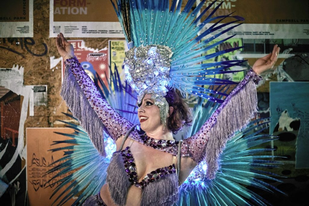 Promotional image of a performer wearing a sparkling lavender costume with fringe detailing and an elaborate blue feathered headdress. The performer stands with arms raised in front of a wall covered with posters and artwork.