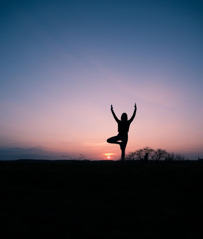 Silhouette of a person balancing in a yoga tree pose at sunset, standing against a pink and blue twilight sky with the sun low on the horizon.
