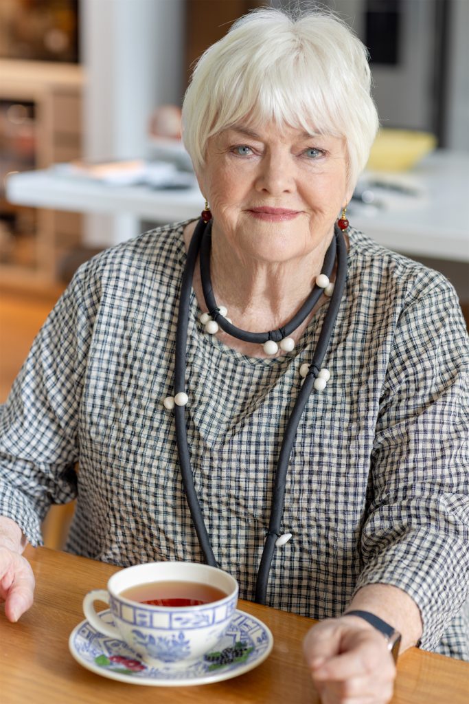 Portrait of Stephanie Alexander seated at a table with a cup of tea in the foreground. The person is wearing a black-and-white checkered top, layered necklace and earrings, with an indoor setting and neutral-toned background softly out of focus.