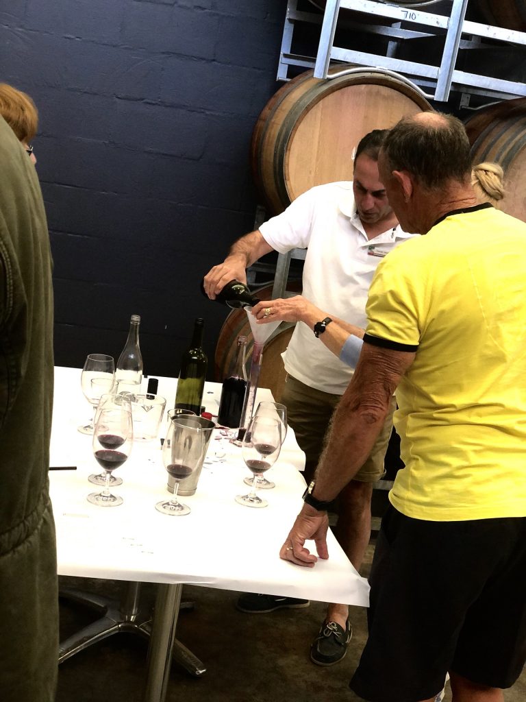 Person demonstrates wine measuring and tasting at a winery table surrounded by wine glasses, bottles, and large wooden barrels.