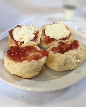 Close-up of four split scones served on a white plate, topped with generous spoonfuls of strawberry jam and cream. The image has a shallow depth of field, with the scones in focus against a soft, blurred background.