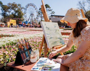 Person paints at an easel outdoors with a ferris wheel, flower beds, and festival attractions behind.
