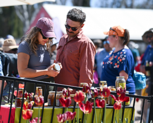 Two people stand at a market stall looking at jars of food products, with red tulips in the foreground and a crowd of visitors in the background.
