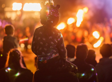 Child seated on an adult’s shoulders watching a fire performance, with flames and a crowd in the background at night.
