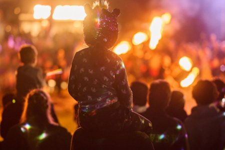 Child seated on an adult’s shoulders watching a fire performance, with flames and a crowd in the background at night.