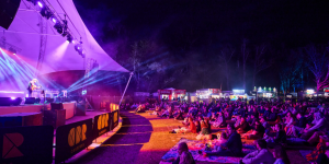 Crowd seated on grass and picnic rugs watching a live performance on a lit stage, with food stalls and trees visible behind.