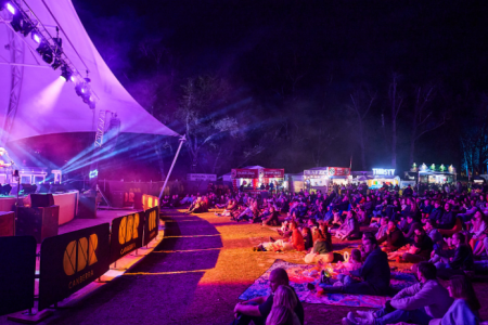 Crowd seated on grass and picnic rugs watching a live performance on a lit stage, with food stalls and trees visible behind.