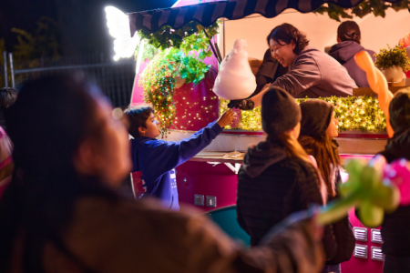 Child reaching up to receive a large piece of fairy floss from a vendor at a brightly lit food stall at night.