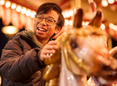 Child riding a carousel horse, holding onto the pole with carousel lights glowing in the background.