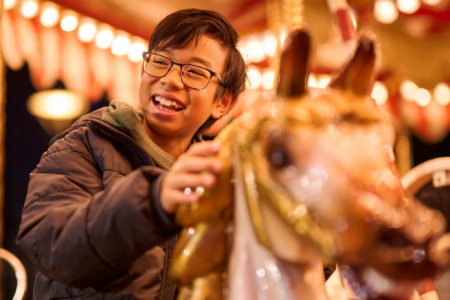 Child riding a carousel horse, holding onto the pole with carousel lights glowing in the background.
