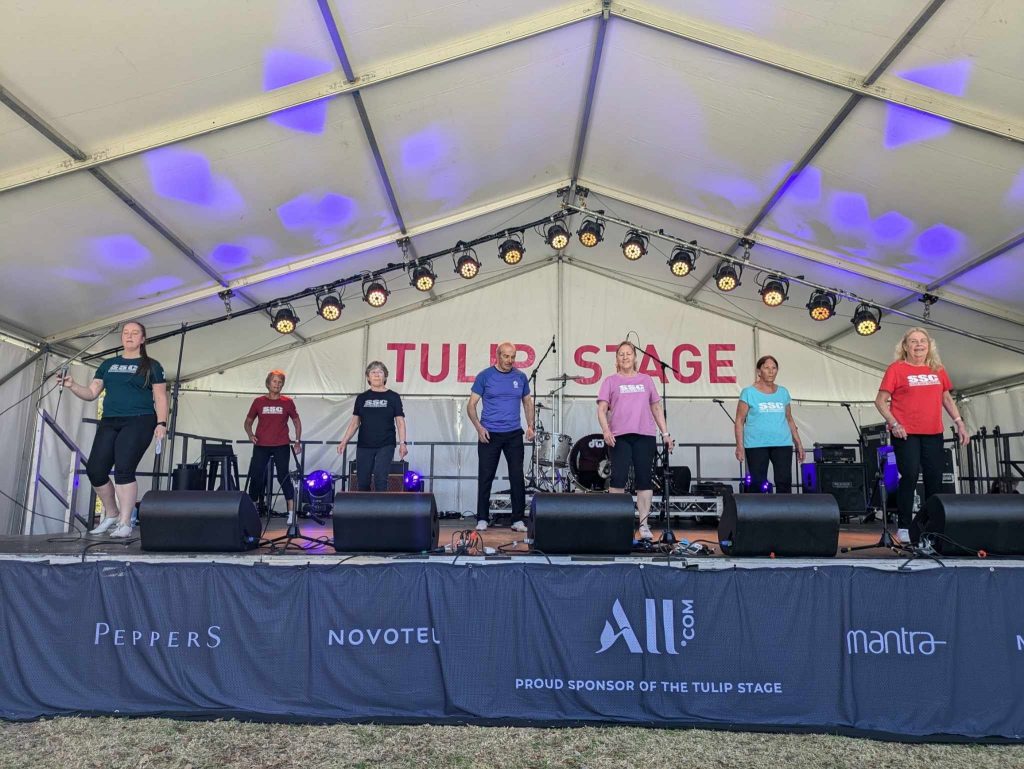 Seven people in colourful SSC shirts perform on the Tulip Stage beneath a large marquee, with stage lights, musical equipment and sponsor signage visible in the background.