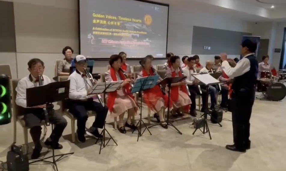 A seated musical ensemble performs indoors while a conductor stands in front leading the group. The performers are arranged in rows with music stands, holding wind instruments such as flutes and clarinets, and wearing coordinated outfits with light tops and red scarves. A drum set is visible toward the back right. Behind the group, a large screen displays event information with the title “Golden Voices, Timeless Hearts” and additional text in English and Chinese. The setting appears to be a community hall or function room with neutral walls and lighting.
