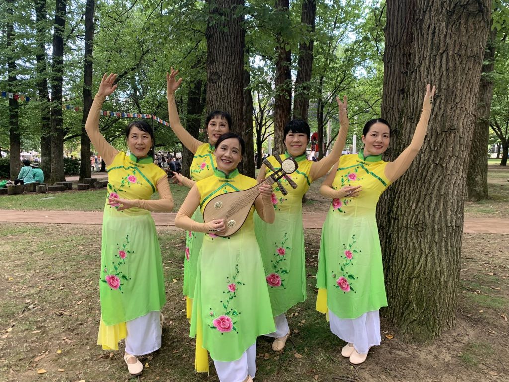 Five performers stand in a park among tall trees, wearing matching green dresses with floral designs and raising their arms in a dance pose. One performer in the center holds a traditional string instrument, while a path, bunting, and people are visible in the background.
