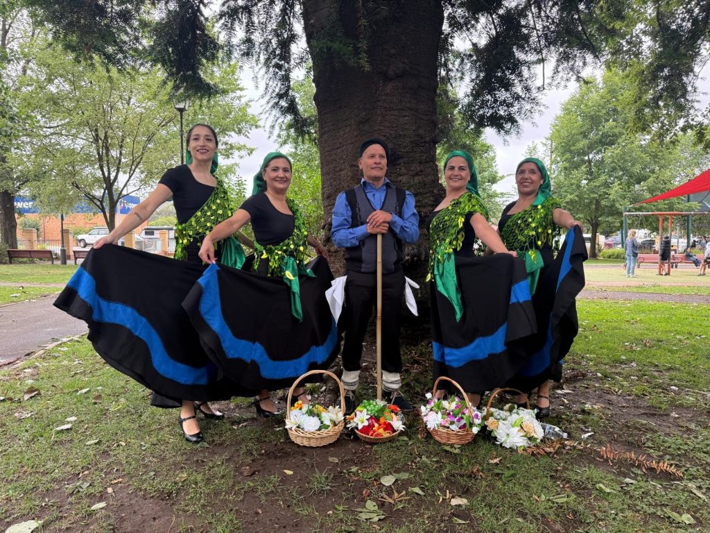 Five performers pose outdoors under a large tree, wearing traditional-style costumes. Four hold up black skirts with blue trim and green decorative tops, while one at center holds a staff. Baskets of flowers and produce are arranged on the ground in front, with a park setting in the background.