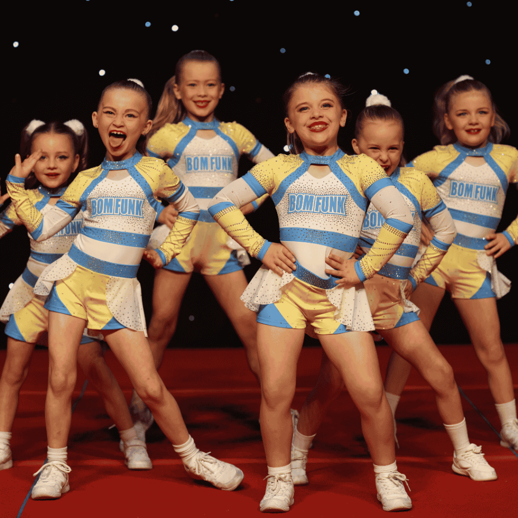 A group of young dancers performs on stage in coordinated yellow, white, and blue costumes with “BOM FUNK” printed across the front. The dancers stand in formation with hands on hips and legs apart, wearing white sneakers against a dark stage background with small star-like lights.