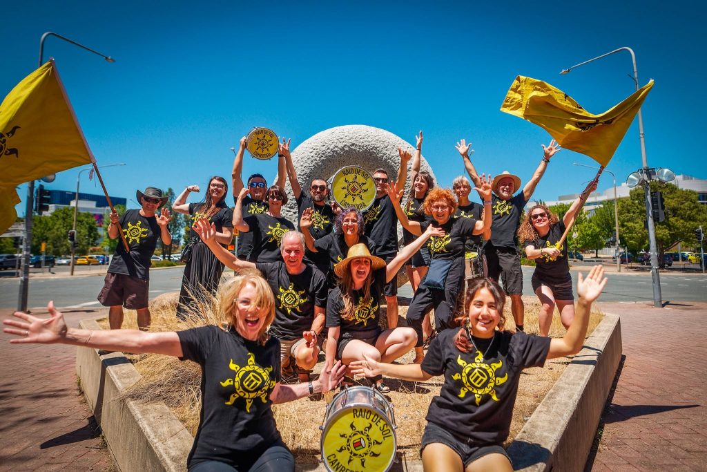 A group of people in matching black shirts play drums and pose with raised arms and yellow flags in a sunny outdoor setting around a large stone sculpture.