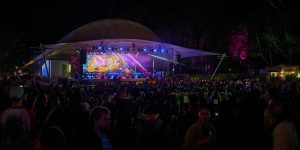 Crowd gathered at an outdoor nighttime concert, facing a large illuminated stage under a canopy structure, with colourful lights and a bright screen displaying “Josh Pyke,” surrounded by trees lit in pink and purple hues.