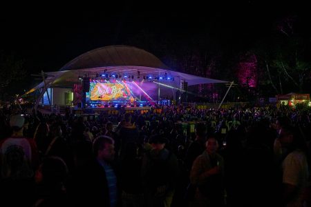 Crowd gathered at an outdoor nighttime concert, facing a large illuminated stage under a canopy structure, with colourful lights and a bright screen displaying “Josh Pyke,” surrounded by trees lit in pink and purple hues.