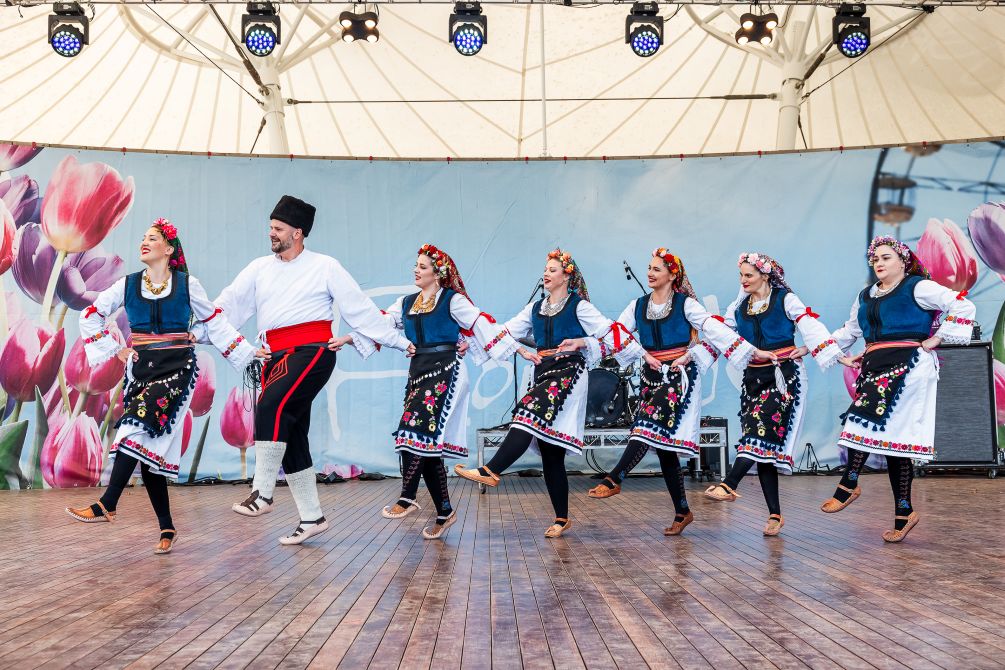 A line of performers in traditional folk costumes dance hand-in-hand on an outdoor stage beneath a canopy, with a backdrop featuring large tulip images.