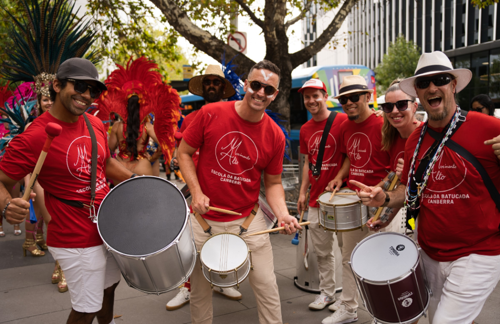 A group of performers in matching red shirts play drums and pose together on a city street, with colourful parade costumes and spectators visible in the background.