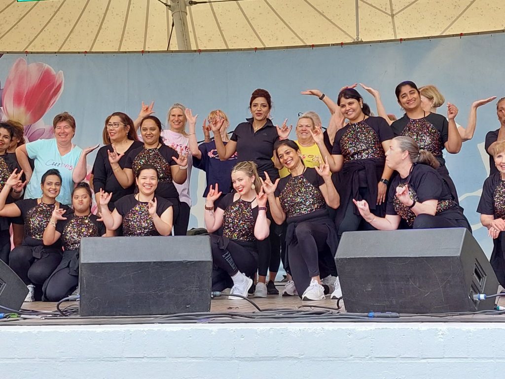 A group of performers in black outfits with sequined tops pose together on an outdoor stage, making hand gestures in front of a backdrop featuring large flower imagery.