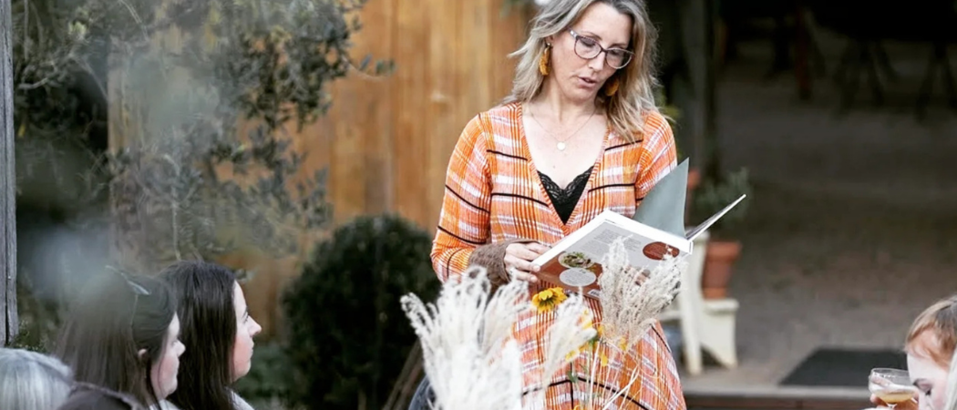 A person in a floral dress stands beside a long outdoor dining table decorated with flowers and candles, while guests gather around in a garden setting.