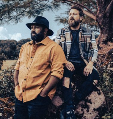Two musicians stand outdoors beneath trees, looking into the distance in a stylised portrait with warm, muted tones.