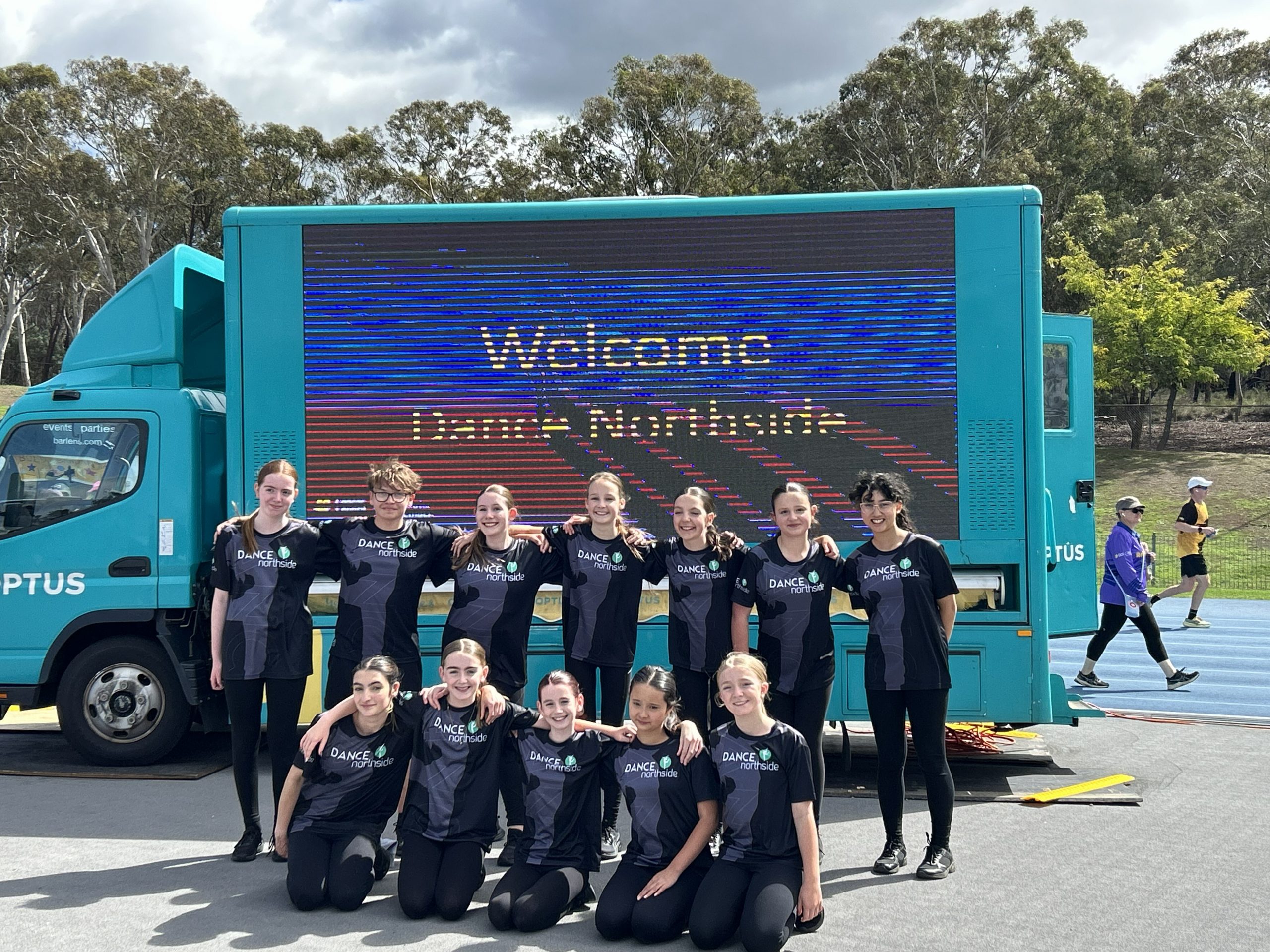 A group of dancers in matching Dance Northside uniforms pose in front of a large mobile screen displaying a welcome message at an outdoor sporting venue.
