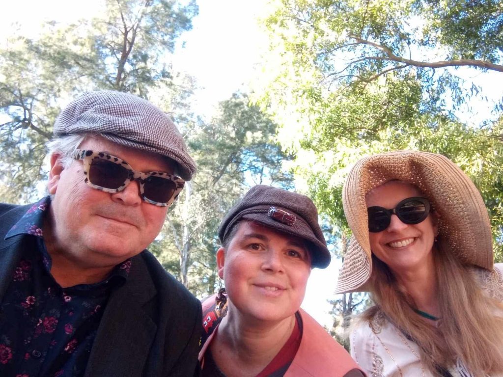 Three people wearing hats and sunglasses pose outdoors in a park, with trees and sunlight in the background.
