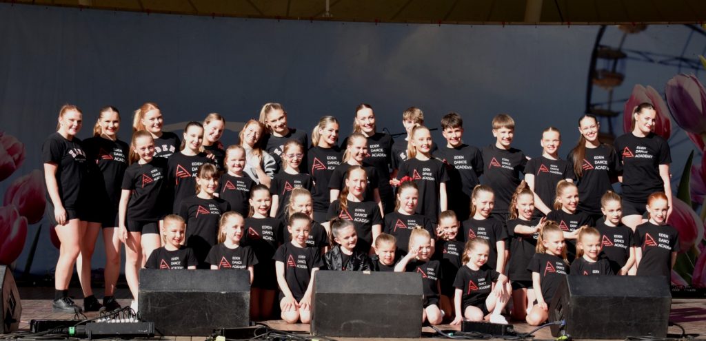 A group of dancers from Eden Dance Academy pose together on an outdoor stage wearing matching black performance shirts in front of a floral backdrop.
