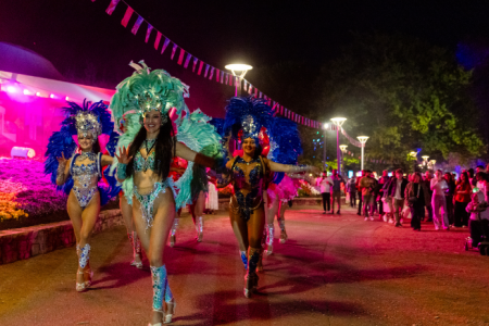Performers in illuminated costumes parade through a crowd beneath colourful festival lights at night.