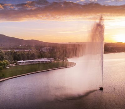 A fountain rises from a lake at sunset, with Canberra's landscape and distant hills in the background.