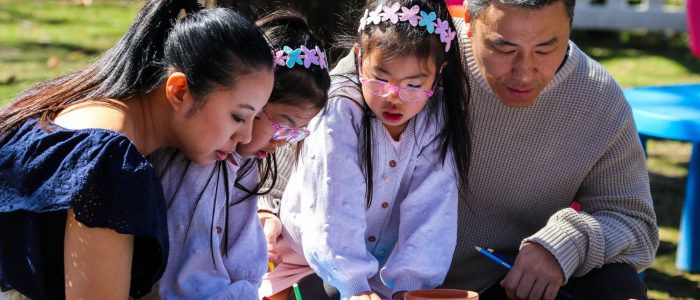 Two adults and three children sit around a small outdoor table, drawing and colouring on activity sheets. The children wear flower headbands and glasses, with coloured pencils and a small terracotta pot on the table in a sunny garden setting.