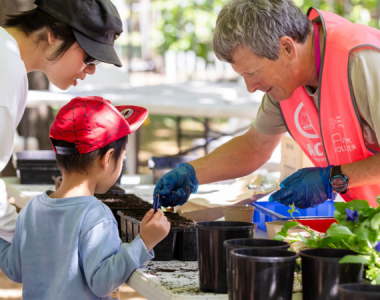 An adult volunteer wearing a bright pink vest helps a child plant seedlings at an outdoor gardening activity, while another adult looks on. Pots, soil, and leafy plants are arranged across a table in a sunlit garden setting.