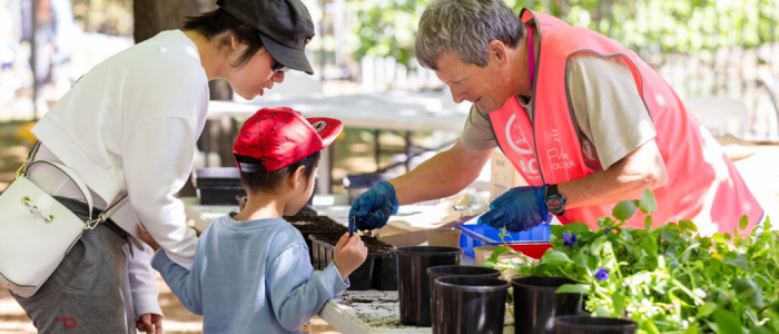 An adult volunteer wearing a bright pink vest helps a child plant seedlings at an outdoor gardening activity, while another adult looks on. Pots, soil, and leafy plants are arranged across a table in a sunlit garden setting.