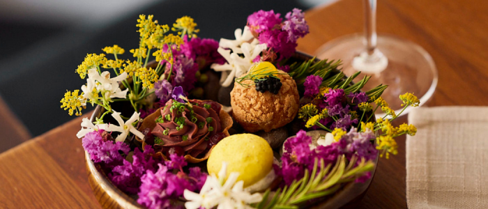 Assorted bite-sized desserts and sweet treats are arranged among colourful edible flowers on a serving dish, accompanied by a glass of white wine on a wooden table.