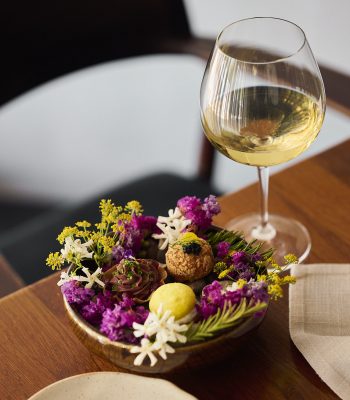 Assorted bite-sized desserts and sweet treats are arranged among colourful edible flowers on a serving dish, accompanied by a glass of white wine on a wooden table.