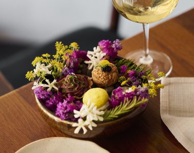 Assorted bite-sized desserts and sweet treats are arranged among colourful edible flowers on a serving dish, accompanied by a glass of white wine on a wooden table.