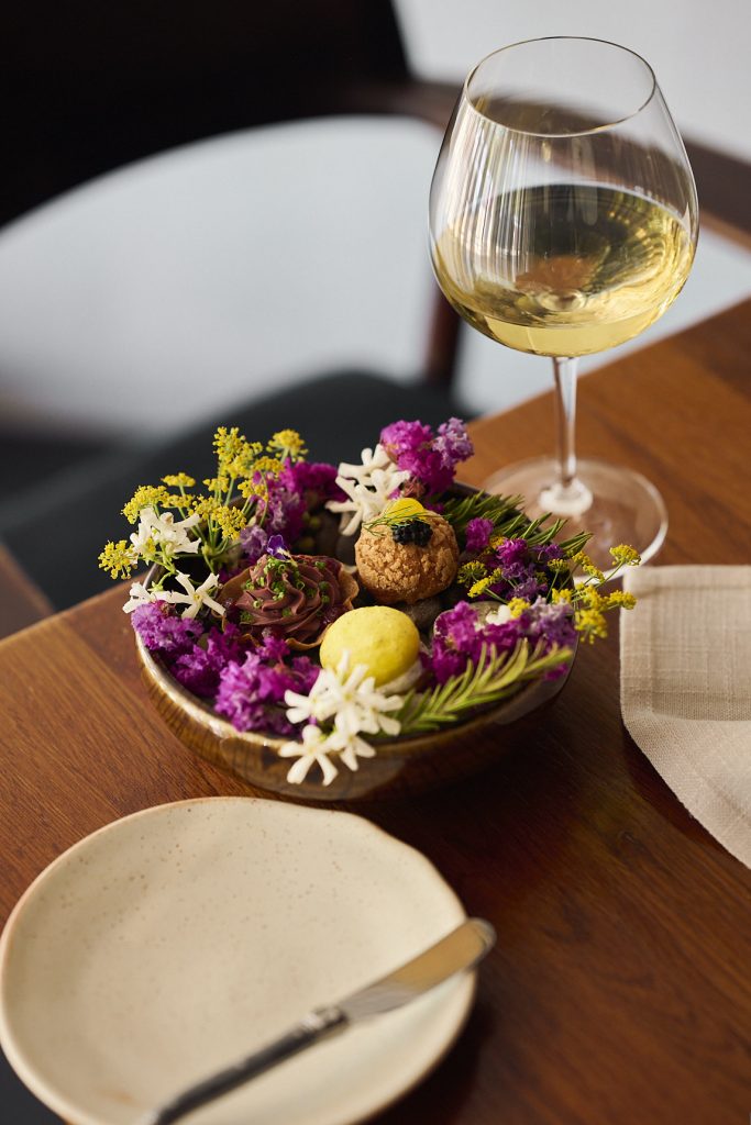 A floral centrepiece featuring purple, yellow, and cream blooms arranged on a dining table beside a glass of white wine.