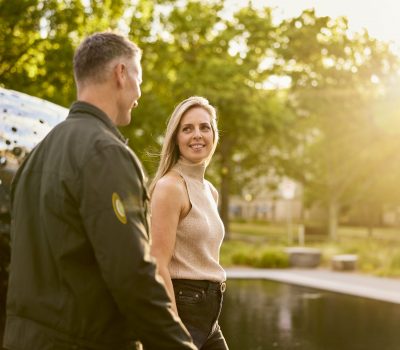 Two people walk beside a reflective water feature in a park at golden hour, with trees, warm sunlight, and a fountain visible in the background.