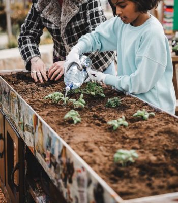 A child and a woman in a hat are planting seeds in a raised garden bed