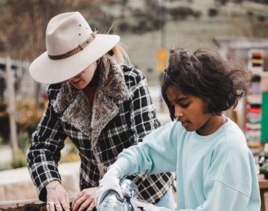 A child and a woman in a hat are planting seeds in a raised garden bed