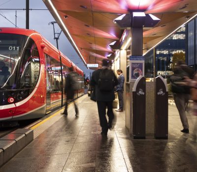 A red light rail tram stops at a city platform at dusk, with passengers walking along the illuminated station.