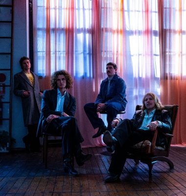 A four-piece band poses for a portrait, featuring the musicians in casual and vintage-inspired clothing against studio backdrop.
