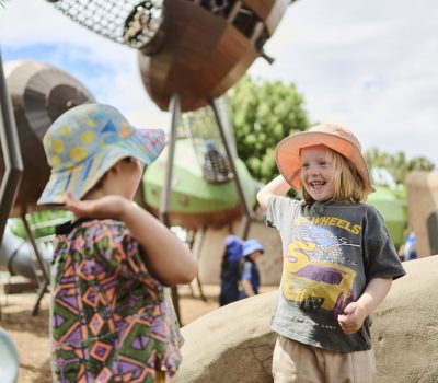 Two children wearing colourful hats explore an outdoor play area featuring large climbing and interactive structures.