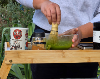 A person whisks vibrant green matcha in a glass bowl on a wooden table, with tea-making accessories and ingredients arranged nearby.