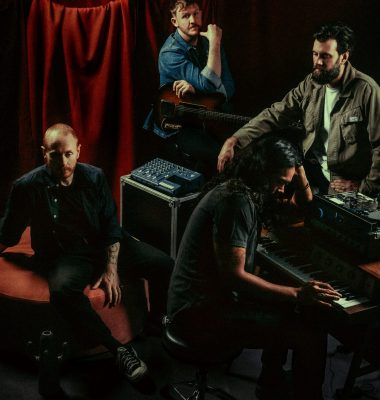 A four-piece band gathers in a dimly lit studio with musical equipment and red curtains in the background.