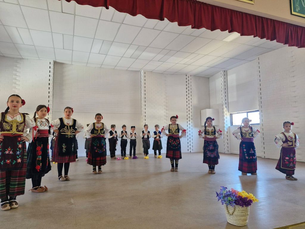 Children in traditional folk costumes performing a group dance on an indoor stage, arranged in rows beneath a red stage curtain.