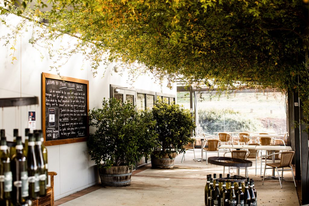 A covered winery terrace features outdoor tables and chairs beneath cascading greenery. Wine bottles are displayed in the foreground, and a chalkboard welcome sign hangs on the wall.

